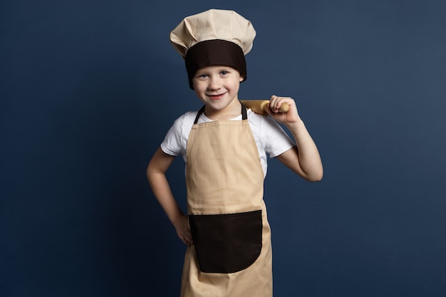 Bild des fröhlichen blauäugigen 7-jährigen Jungenkochs in der Kochuniform, der Nudelholz auf seiner Schulter hält, froh ist, während Teig für Lebkuchenplätzchen knetet, Kamera mit glücklichem Lächeln betrachtend