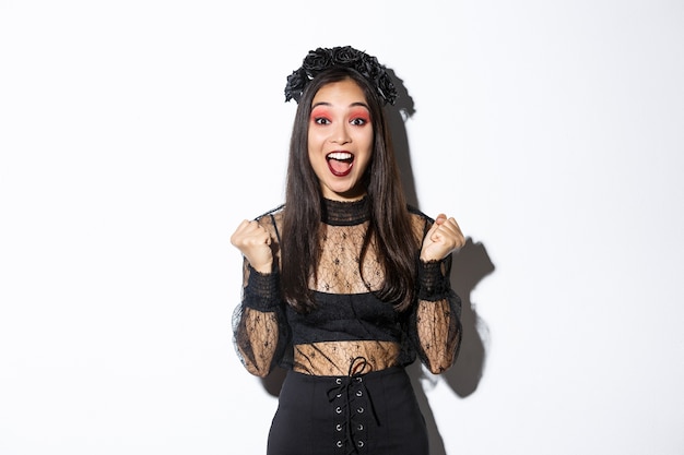 Kostenloses Foto bild des glücklichen asiatischen gotischen mädchens im halloween-partykleid, das sich über den sieg freut, triumphiert und ja sagt und über dem weißen hintergrund steht, der den sieg feiert.