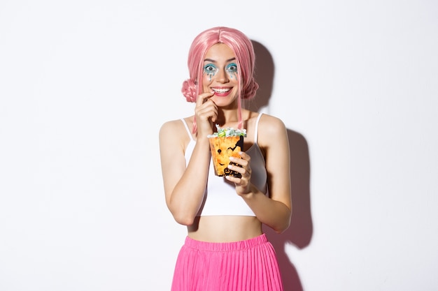 Bild des glücklichen attraktiven Mädchens mit rosa Perücke und hellem Make-up, das Süßes oder Saures geht, Halloween feiert, Süßigkeiten zeigt und lächelt, stehend.