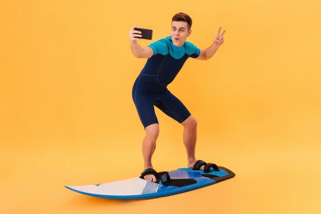 Bild des glücklichen Surfers im Neoprenanzug, der Surfbrett wie auf Welle verwendet, während Selfie auf Smartphone macht und Friedensgeste zeigt