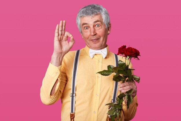 Bild des gutaussehenden älteren Mannes, der okay Zeichen zeigt, bereit, weiter zu datieren, hält rote Blumen in der Hand, trägt gelbes Hemd und Fliege, lokalisiert auf rosa, posierend im Studio. Körpersprachenkonzept.