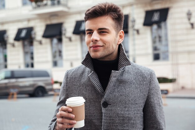 Bild des gutaussehenden Mannes Mitnehmerkaffee von der Papierschale genießend, beim Gehen hinunter leere Straße