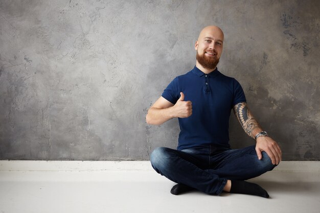 Bild des gutaussehenden positiven jungen kahlen bärtigen Kerls mit Tätowierung auf seinem muskulösen Arm, der breit lächelt und Daumen hoch Geste macht, großen Tag hat, in guter Stimmung ist, Zustimmung zeigt