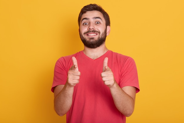 Bild des hübschen lächelnden modernen Mannes kleidet rotes lässiges T-Shirt, das ok Zeichen mit beiden Daumen zeigt, Modell posiert isoliert auf gelbem, bärtigem jungem Mann mit glücklichem Gesichtsausdruck.