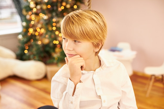 Bild des hübschen Teenagers im weißen Hemd mit nachdenklichem nachdenklichem Blick, Kinn berührend, denkend, wo Mutter Neujahrsgeschenke versteckte, im Wohnzimmer mit Weihnachtsbaum posierend