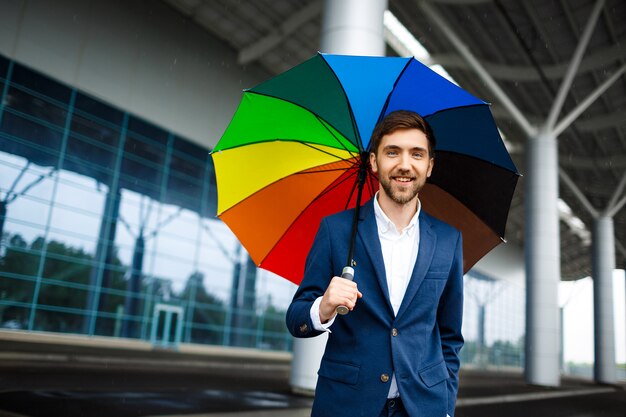 Bild des jungen fröhlichen Geschäftsmannes, der bunten Regenschirm in der regnerischen Straße hält