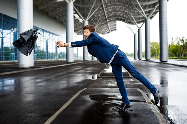 Bild des jungen Geschäftsmannes auf regnerischem Flughafen, der gebrochenen Regenschirm fängt