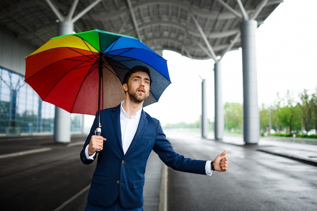 Bild des jungen Geschäftsmannes, der bunten Regenschirm hält Auto am Terminal hält