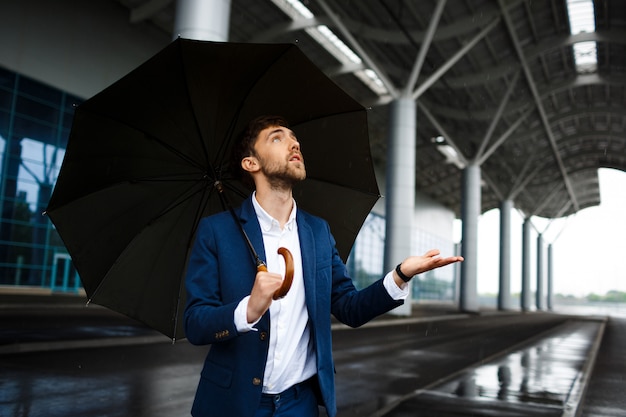 Bild des jungen Geschäftsmannes, der Regenschirm im Regen hält, der Tropfen betrachtet
