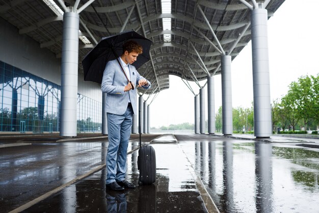Bild des jungen rothaarigen Geschäftsmannes, der schwarzen Regenschirm im Regen hält und auf Uhr schaut