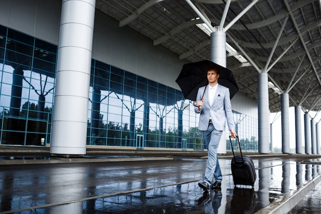 Bild des jungen rothaarigen Geschäftsmannes, der schwarzen Regenschirm und Koffer hält, die im Regen am Bahnhof gehen