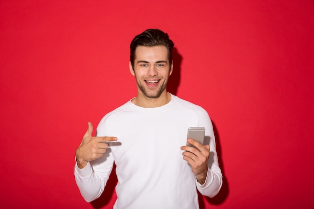 Bild des lächelnden Mannes in der schauenden Strickjacke beim Halten von Smartphone und Zeigen auf ihn über roter Wand