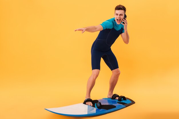 Bild des lächelnden Surfers im Neoprenanzug unter Verwendung des Surfbretts beim Sprechen durch Smartphone