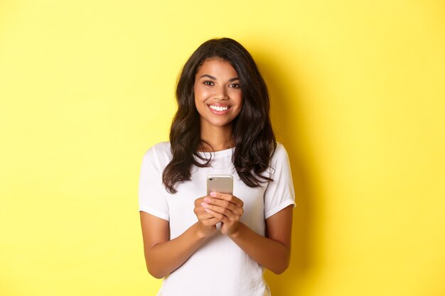 Bild des modernen afroamerikanischen Mädchens, das mit dem Handy lächelt, das über gelbem Hintergrund steht