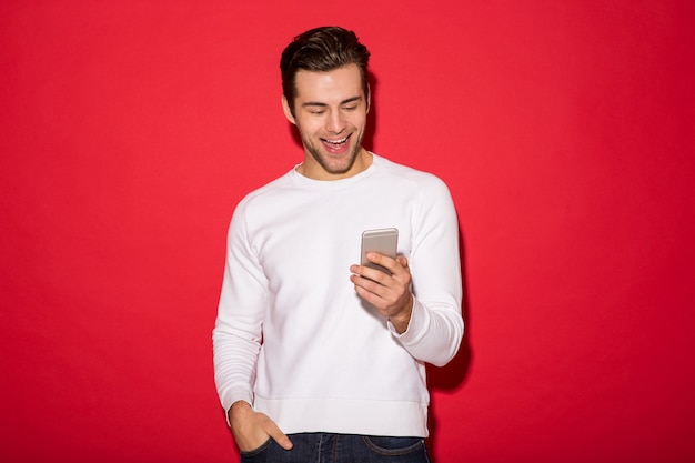 Bild des netten Mannes in der Strickjacke unter Verwendung des Smartphone über roter Wand