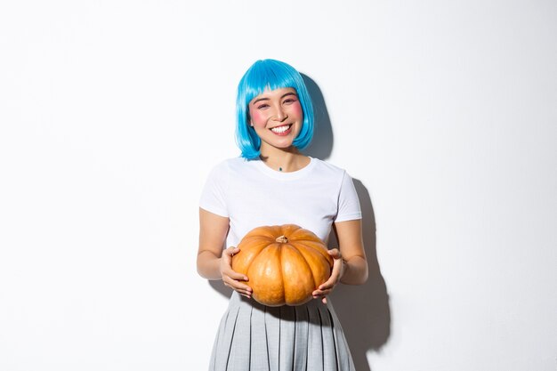 Bild des niedlichen asiatischen Mädchens, das Ihnen Kürbis für Halloween-Partei gibt, blaue Perücke tragend, stehend.