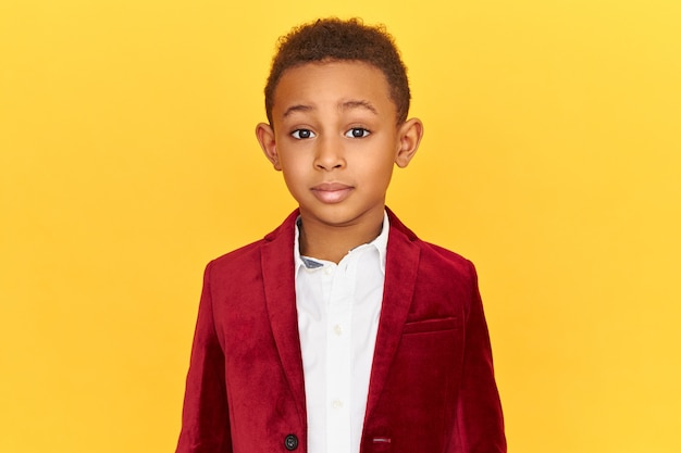 Bild des niedlichen charmanten kleinen afroamerikanischen Jungen, der stilvolle Kleidung trägt, die isoliert betrachtet Kamera in der Faszination aufwirft, erstaunt mit großen Verkaufspreisen