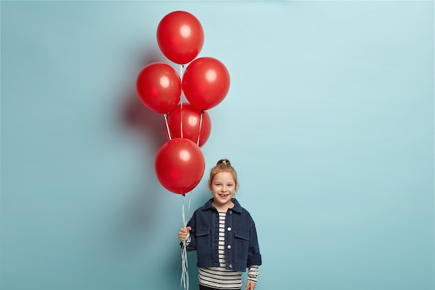 Bild des niedlichen kleinen Kindes in der modischen Jeansjacke steht mit roten Luftballons, kommt am Geburtstag des Freundes, hat glücklichen Gesichtsausdruck, steht über blauer Wand. Kindheits- und Feierkonzept