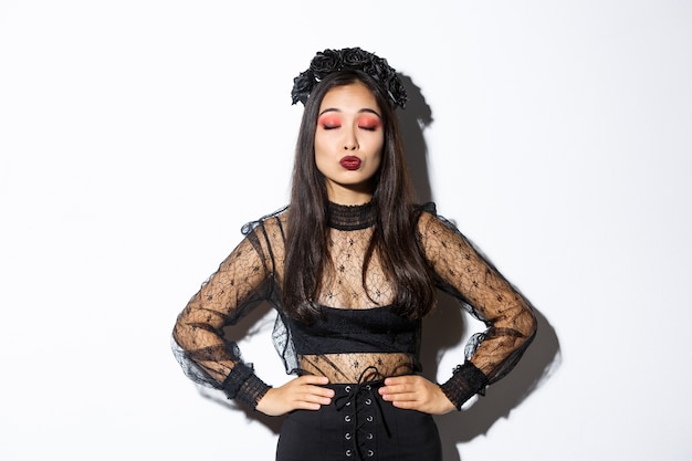 Bild des schönen asiatischen Mädchens im Halloween-Kostüm schließen Augen und schmollen, warten auf Kuss, stehend über weißem Hintergrund, der gotisches schwarzes Kleid mit Kranz aus Rosen macht.