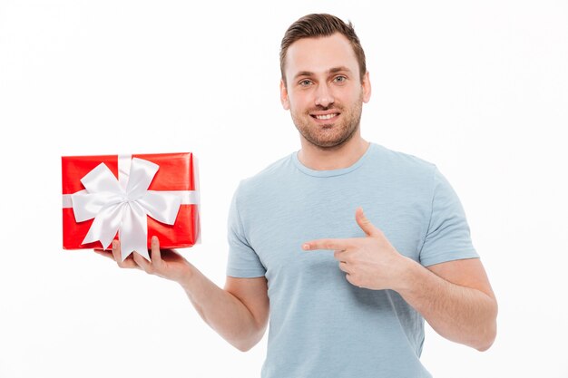 Bild des schönen Kerls, der die Borste lächelt und Zeigefinger auf roter Geschenkbox zeigt