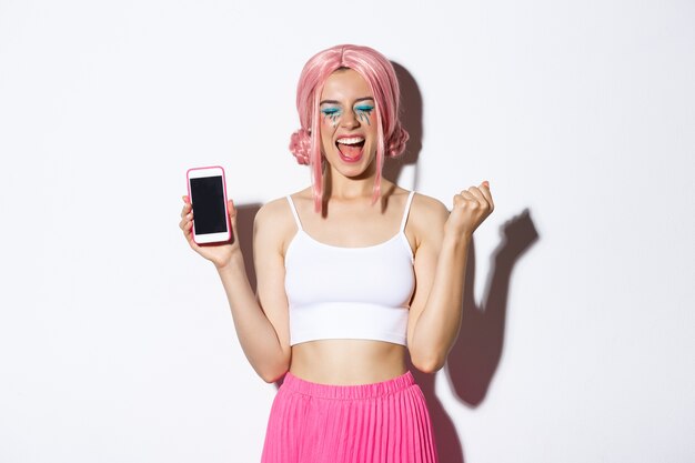 Bild des schönen Partygirls, das sich freut, Feiertag feiert, Smartphonebildschirm zeigt und vor Freude schreit, rosa Perücke und Halloween-Outfit tragend.