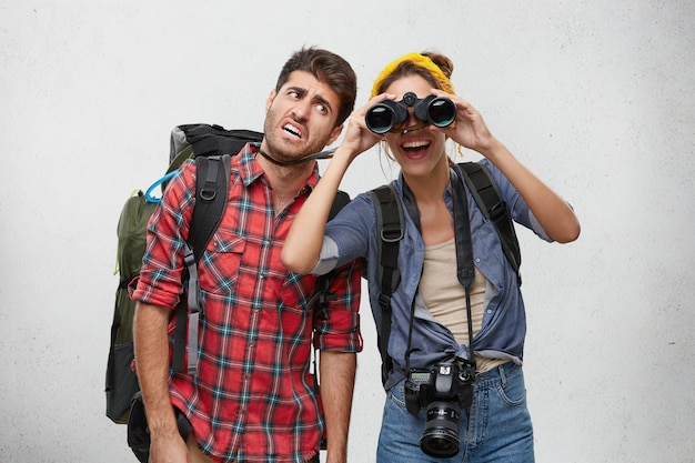 Bild des touristen des müden bärtigen mannes, der schweren rucksack und fröhliche aufgeregte frau mit fotokamera trägt, die nach platz für camping unter verwendung des fernglases während der wanderreise zusammen sucht. menschen und abenteuer