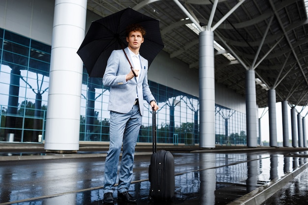 Bild des zuversichtlichen jungen rothaarigen Geschäftsmannes, der schwarzen Regenschirm und Koffer im Regen am Terminal hält