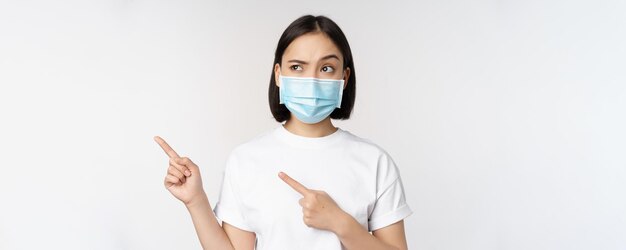 Bild einer asiatischen Frau in medizinischer Maske von Covid, die verwirrt nach links schaut und auf das Logo zeigt, das Werbung zeigt, die auf weißem Hintergrund steht
