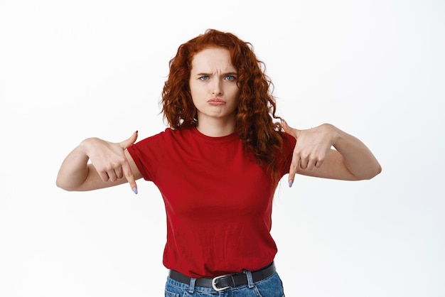 Kostenloses Foto bild einer enttäuschten und traurigen frau, die sich über unfaire dinge beschwert, die schmollen und die stirn runzeln und mit dem finger nach unten auf ein banner zeigen, das vor weißem hintergrund steht