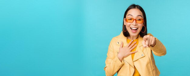 Bild einer glücklichen Koreanerin mit Sonnenbrille, die mit dem Finger auf die Kamera zeigt, mit erstauntem, überraschtem und freudigem Gesichtsausdruck, der über blauem Hintergrund steht