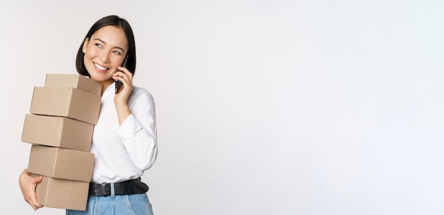 Bild einer jungen asiatischen Geschäftsfrau, die einen Anruf entgegennimmt, während sie Kisten für die Lieferung trägt, die vor weißem Hintergrund posieren