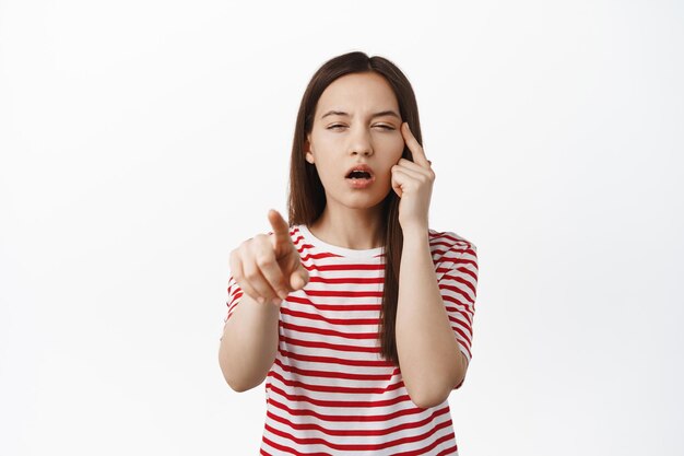 Bild einer jungen Frau, die mit dem Finger zeigt, die Augen ohne Brille zusammenkneift, nichts sehen kann und versucht, mit einer Brille etwas zu lesen, die in einem gestreiften T-Shirt vor weißem Hintergrund steht