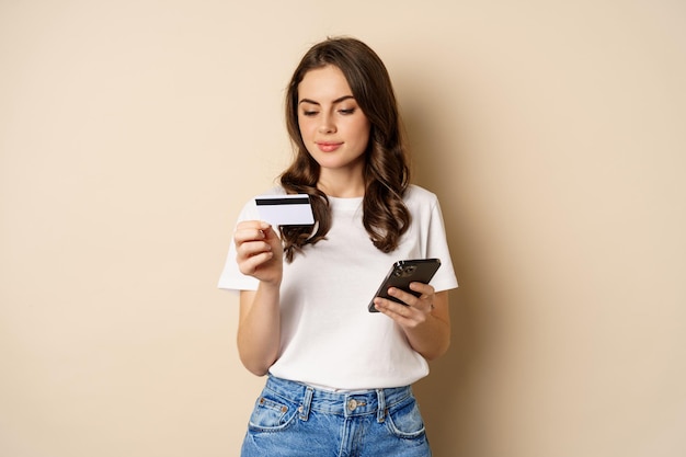 Bild einer jungen glücklichen Frau, die online bezahlt, Smartphone und Kreditkarte in der Hand hält, Informationen in die Anwendung auf dem Mobiltelefon eingibt und vor beigem Hintergrund steht