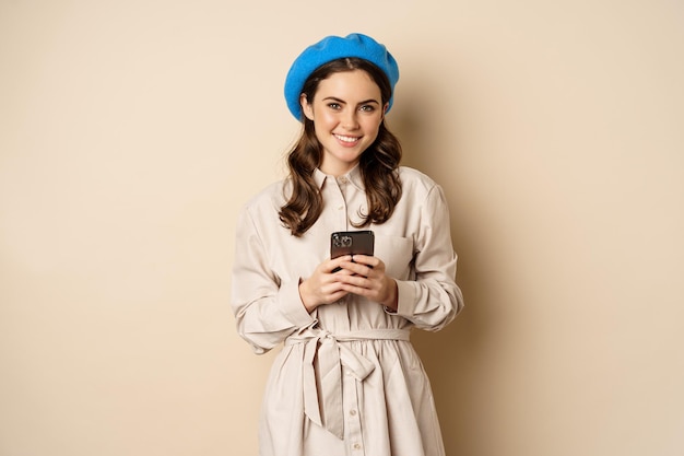 Bild einer jungen stilvollen Französin im Trenchcoat, die mit Smartphone steht und eine Handy-App verwendet ...