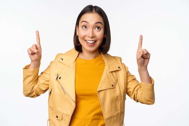 Bild einer lächelnden asiatischen brünetten Frau, die mit dem Finger nach oben zeigt und Werbung mit fröhlichem Gesicht zeigt, das vor weißem Hintergrund posiert