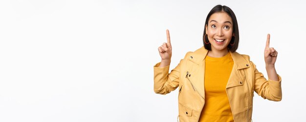 Bild einer lächelnden asiatischen brünetten Frau, die mit dem Finger nach oben zeigt und Werbung mit fröhlichem Gesicht zeigt, das vor weißem Hintergrund posiert