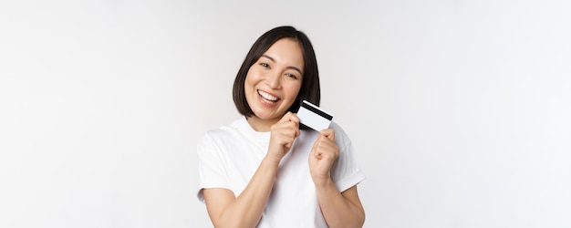 Bild einer lächelnden asiatischen Frau, die eine Kreditkarte umarmt, die kontaktlos im weißen T-Shirt auf weißem Hintergrund kauft