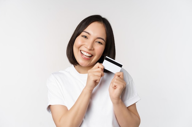 Bild einer lächelnden asiatischen Frau, die eine Kreditkarte umarmt, die kontaktlos im weißen T-Shirt auf weißem Hintergrund steht Kopierplatz