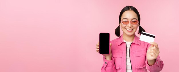 Bild einer lächelnden Koreanerin, die die Schnittstelle der Smartphone-Anwendung mit Kreditkarte und Handybildschirm zeigt, die kontaktlos beim Online-Shopping über rosa Hintergrund steht
