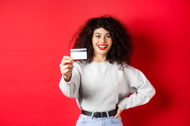Bild einer modernen Frau mit lockigem Haar, Hand ausstrecken und Plastikkreditkarte zeigend, Bank- oder Einkaufsangebot empfehlend, roter Hintergrund.