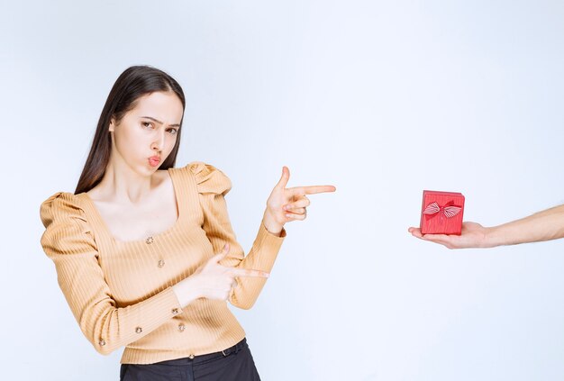 Bild einer netten jungen Frau, die auf eine rote kleine Geschenkbox steht und zeigt.