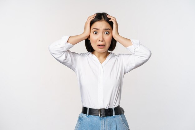 Bild einer schockierten, ängstlichen asiatischen Frau in Panik, die Hände auf dem Kopf hält und sich Sorgen macht, frustriert und verängstigt vor weißem Hintergrund zu stehen