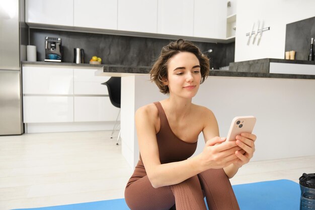 Bild einer schönen Frau in stylischer Sportkleidung, die zu Hause auf einer Yogamatte sitzt und auf ihr Smartphone schaut