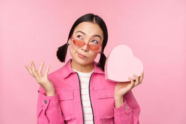 Bild einer stilvollen asiatischen Freundin mit Sonnenbrille, die errät, was sich in der herzförmigen Geschenkbox befindet, die über rosa Hintergrund steht