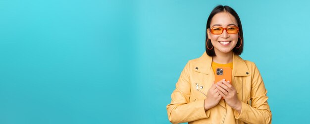 Bild einer stilvollen, schönen Chinesin mit Sonnenbrille, die ein Smartphone hält und ein Foto auf dem Handy macht und lächelnd etwas aufzeichnet, das über blauem Hintergrund steht