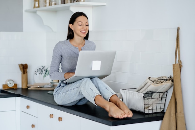 Bild einer süßen brünetten Frau, die auf dem Tisch in der Küche sitzt und einen Laptop benutzt