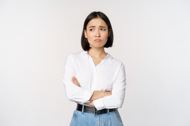 Bild einer traurigen asiatischen Frau im Büro, die schmollend und die Stirn runzelnd enttäuscht stehend verärgert und beunruhigt vor weißem Hintergrund steht