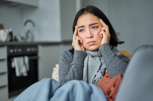 Kostenloses Foto bild einer traurigen jungen frau, die sich krank fühlt, weil sie mit einem schal unwohl sitzt und in ihrem wohnzimmer die stirn runzelt