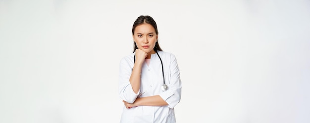 Bild einer wütenden asiatischen Ärztin, die genervt und beunruhigt die Augenbrauen runzelt und nachdenklich vor weißem Hintergrund steht