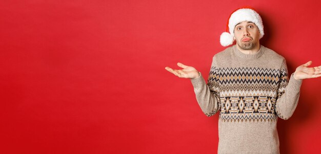 Bild eines ahnungslosen jungen Mannes in Pullover und Weihnachtsmütze, der mit den Schultern zuckt und ahnungslos aussieht, weiß nicht, was er für die Neujahrsferien kaufen soll, und steht auf rotem Hintergrund.